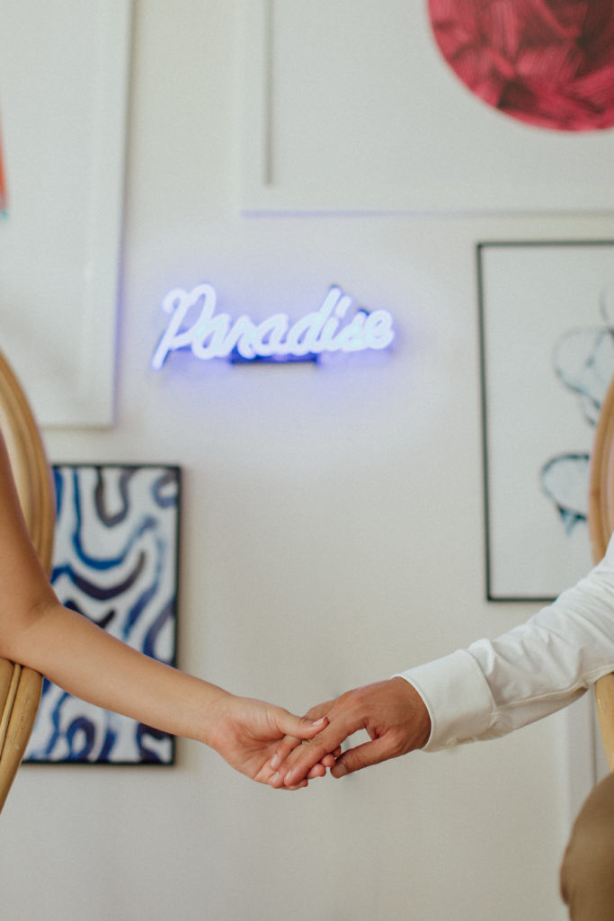 the couple holding hands with the neon sign "Paradise" behind them on the wall